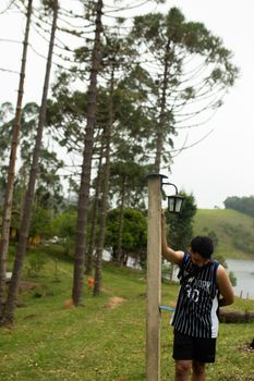 A man casually leans against a lamppost in a serene countryside setting by a lakeside.