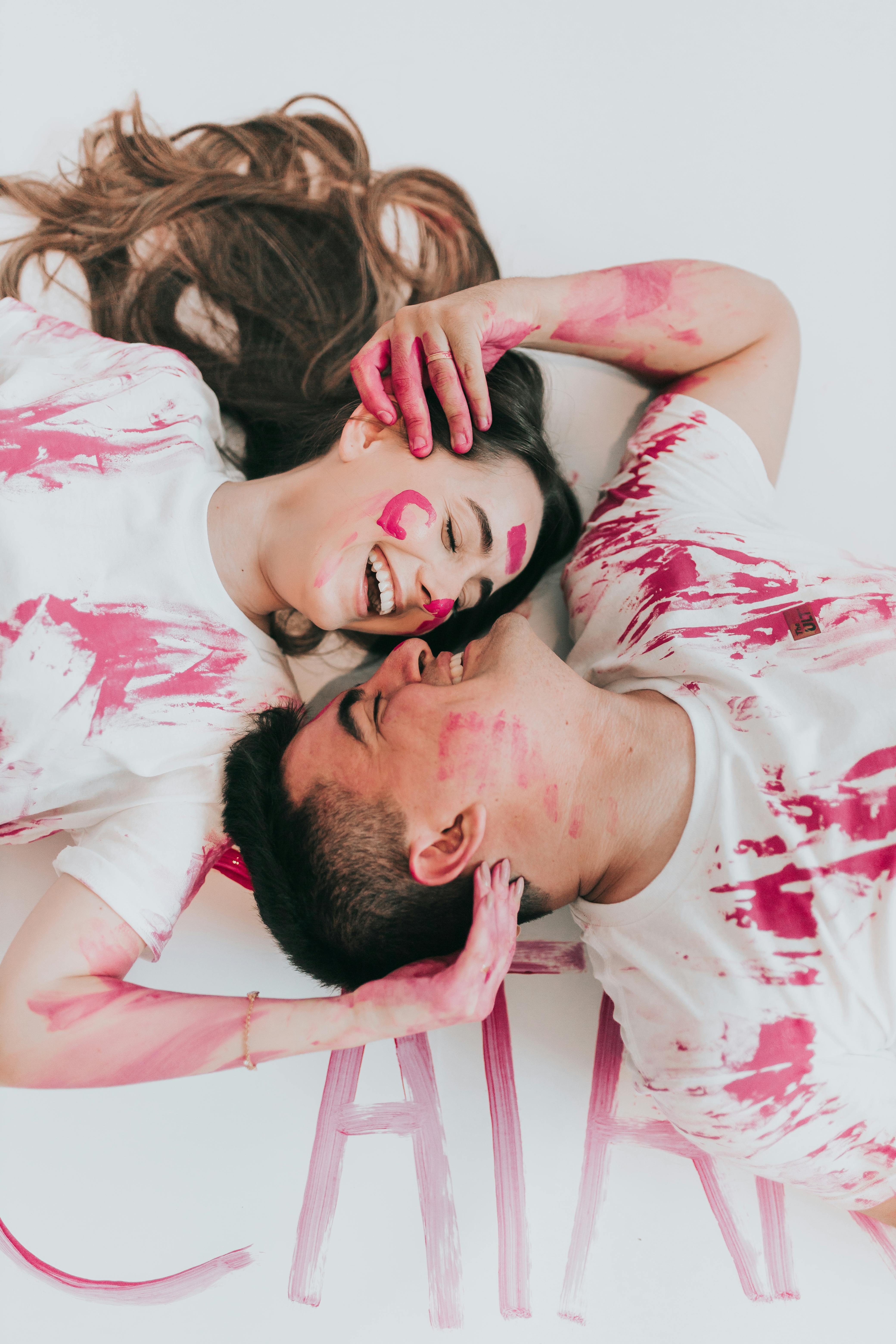 Happy couple lying on floor with playful pink paint splatters on their clothes and faces.