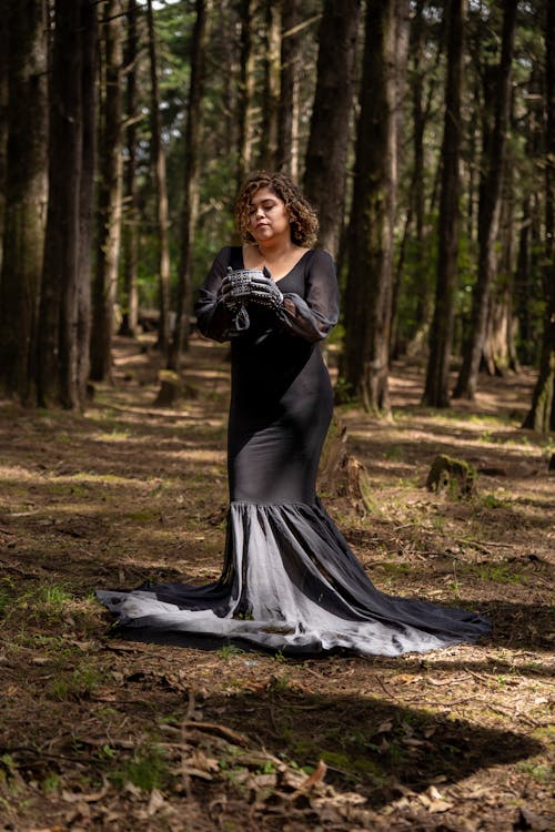 Женщина в длинном черном платье стоит в лесу.