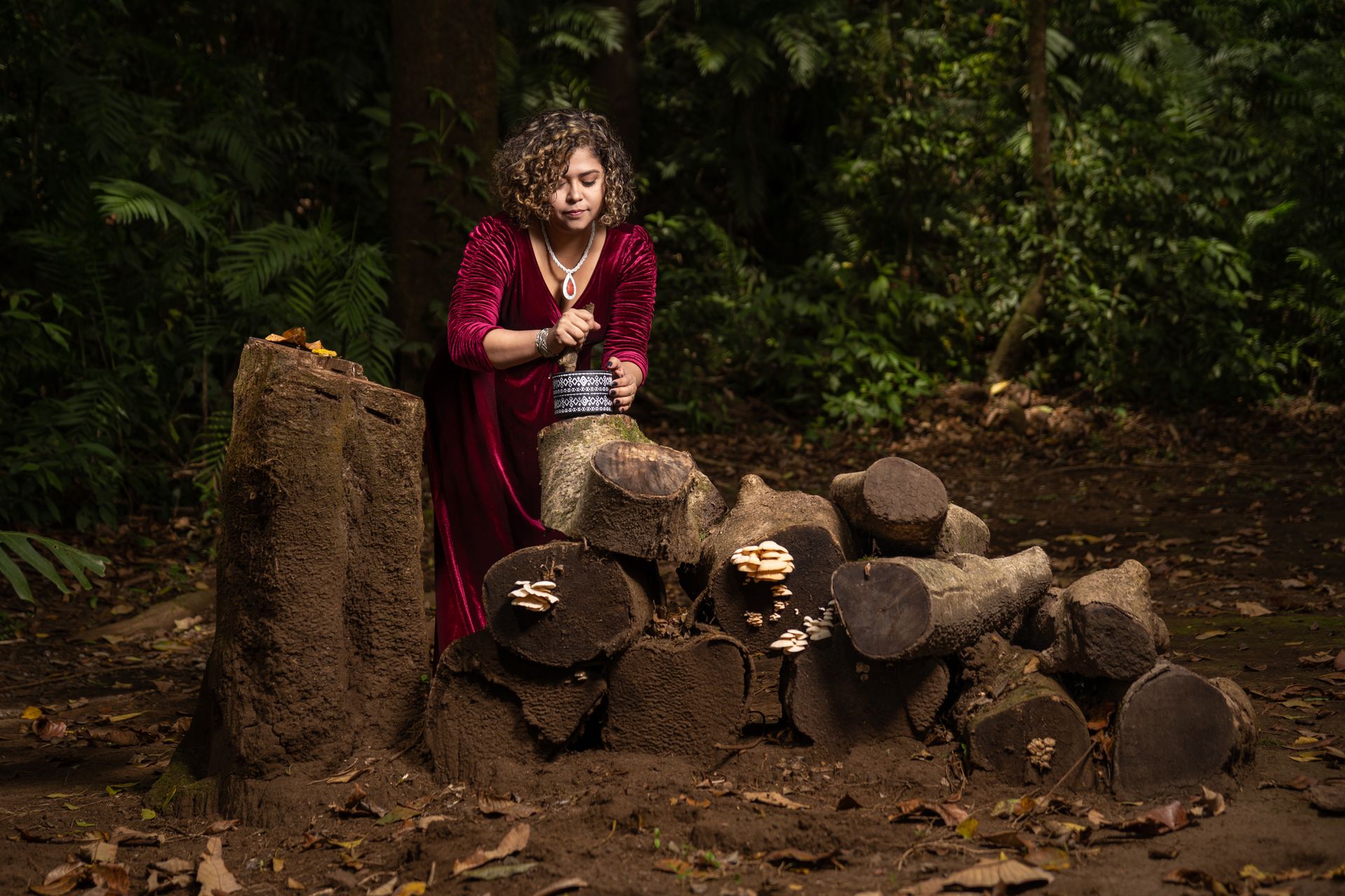 A woman in a red velvet dress stands beside logs, surrounded by lush forest.
