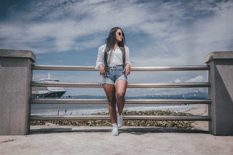 Woman In Shorts And Sunglasses Standing On A Pier On The Background Of Sea And Ship 