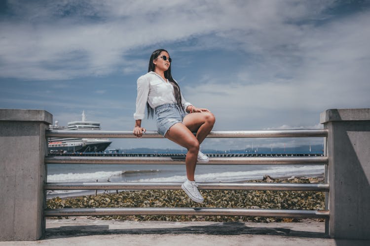 Woman In Shorts And Sunglasses Sitting On A Pier On The Background Of Sea And Ship 