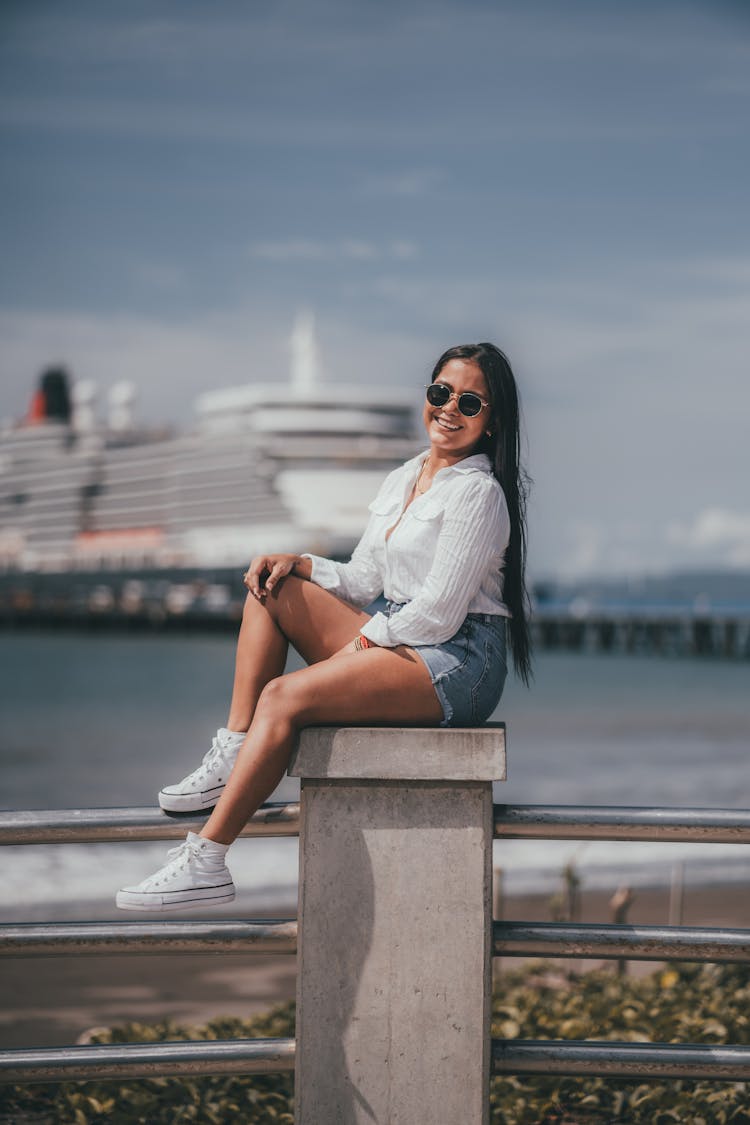 Woman In Shorts And Sunglasses Sitting On A Pier On The Background Of Sea And Ship 