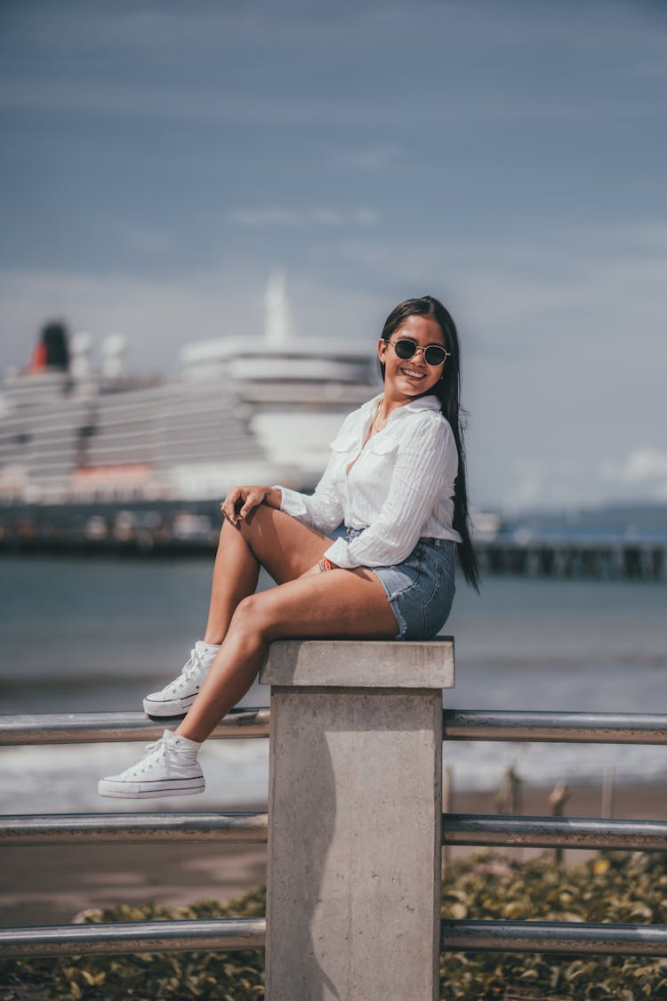 Woman In Shorts And Sunglasses Sitting On A Pier On The Background Of Sea And Ship 