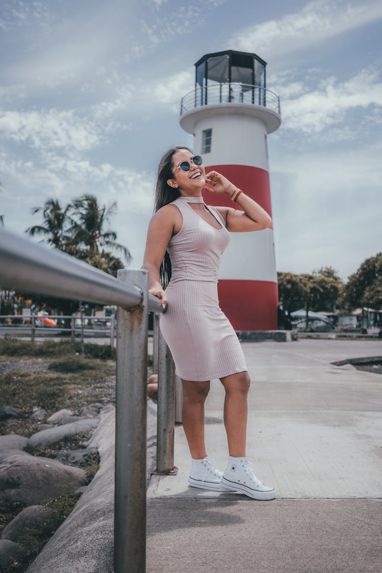 Woman In A Dress And Sunglasses Standing On A Pier Near A Lighthouse 