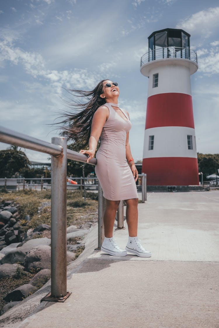 Woman In A Dress And Sunglasses Standing On A Pier Near A Lighthouse 