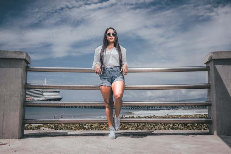 Woman In Shorts And Sunglasses Standing On A Pier On The Background Of Sea And Ship 