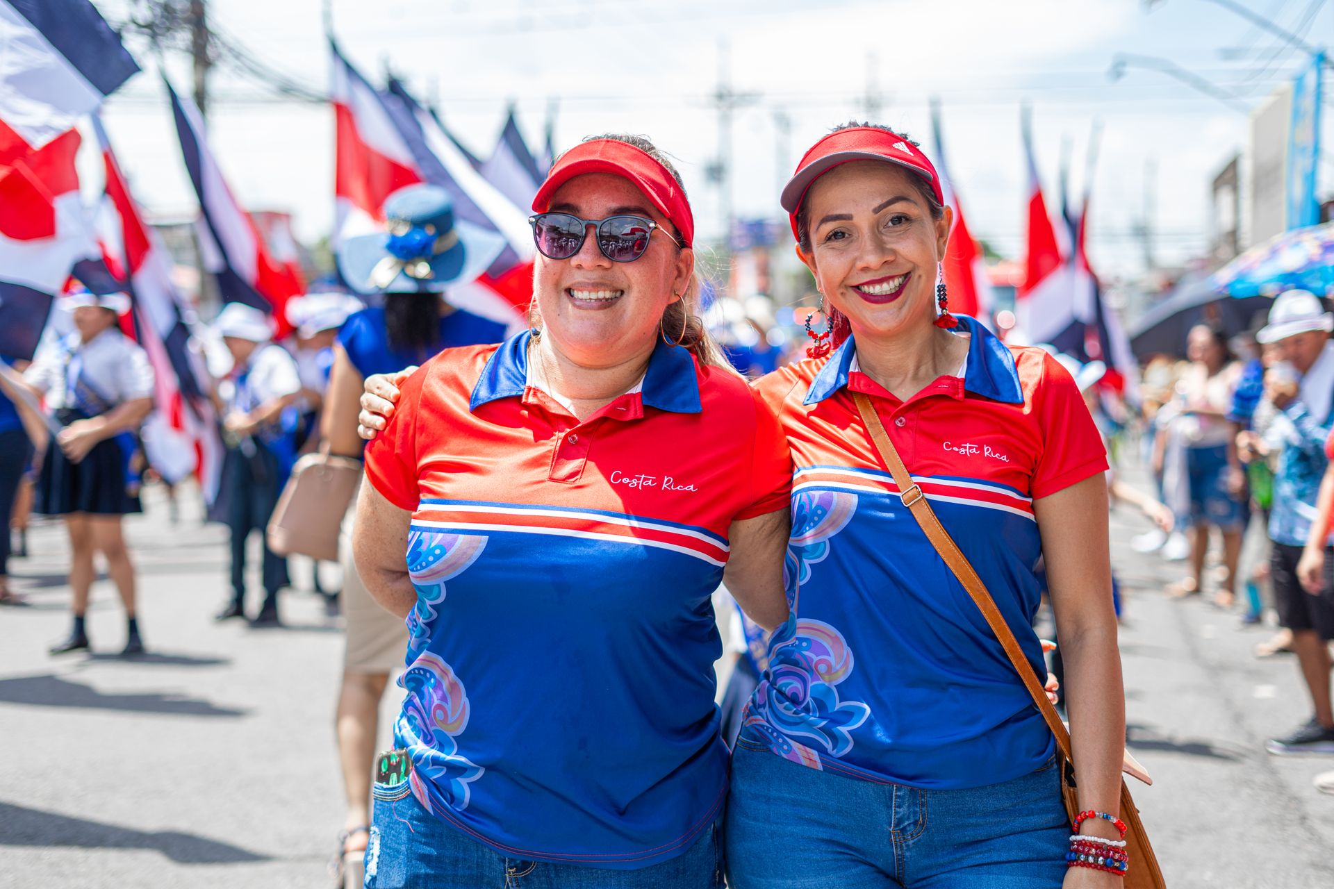 Two women in vibrant shirts celebrate at a festive Costa Rican parade with flags and smiles.