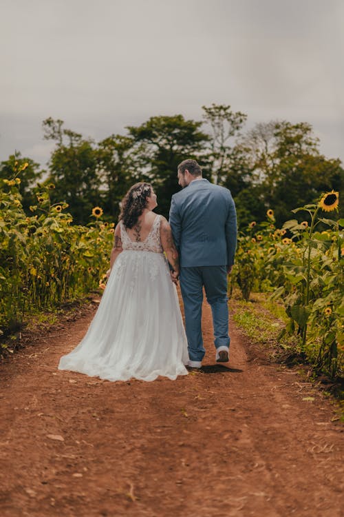 Een Bruid En Bruidegom Lopen Over Een Onverharde Weg In Een Zonnebloemveld