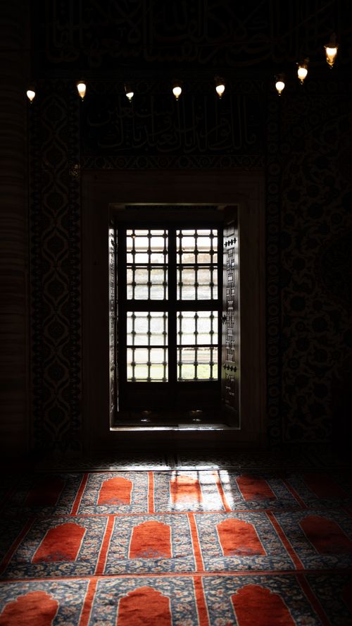 Free Capture of a serene mosque interior featuring a patterned window and red prayer rugs. Stock Photo