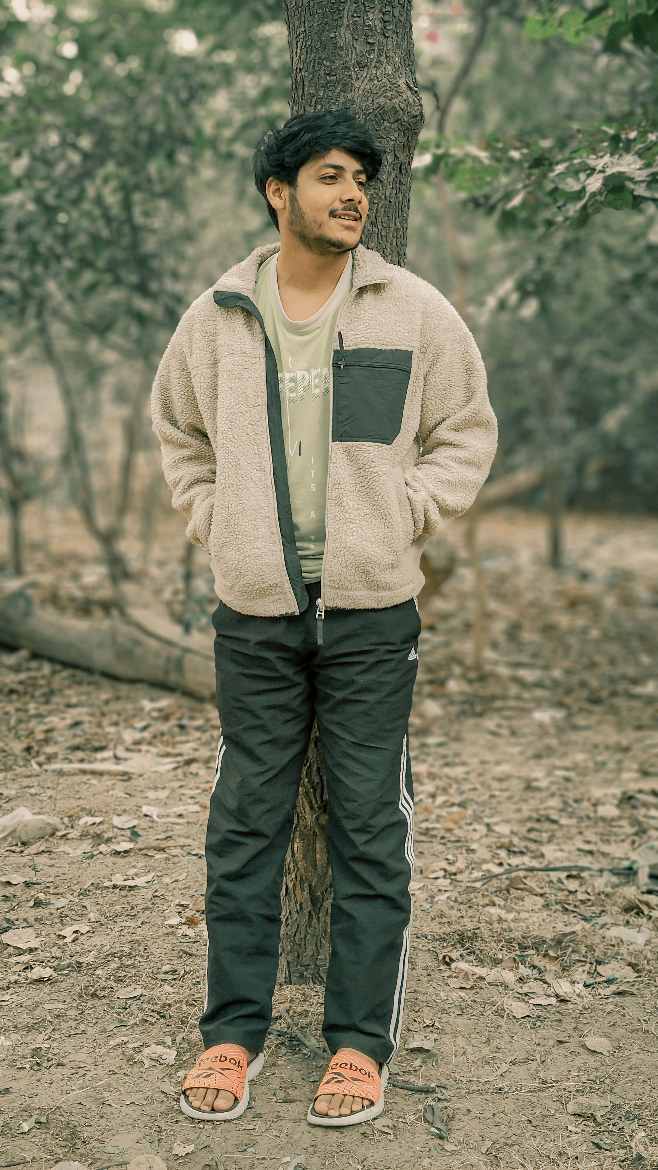 A young man in a sherpa jacket stands relaxed in a quiet forest setting, hands in pockets.