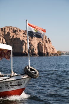 Boat with Egyptian flag on the Nile with rocky outcrop in the background, sunlit scene.