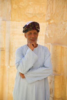 Elderly man in traditional attire poses against ancient stone wall in Luxor, Egypt.