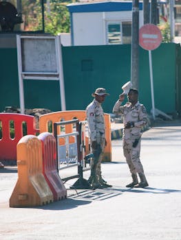 Two soldiers in desert camouflage stand at a checkpoint with barriers outdoors.