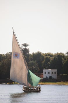 A felucca sails on the Nile River in Luxor, Egypt, with a scenic background of palm trees.