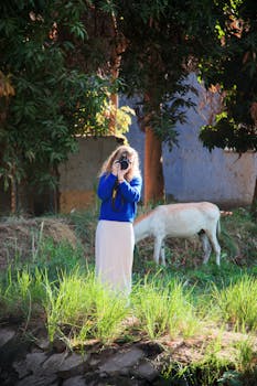 A woman captures outdoor photography amidst vibrant greenery and a goat grazing peacefully.