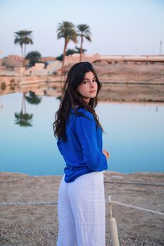 A woman in blue stands beside a tranquil pool reflecting palm trees in Luxor, Egypt.