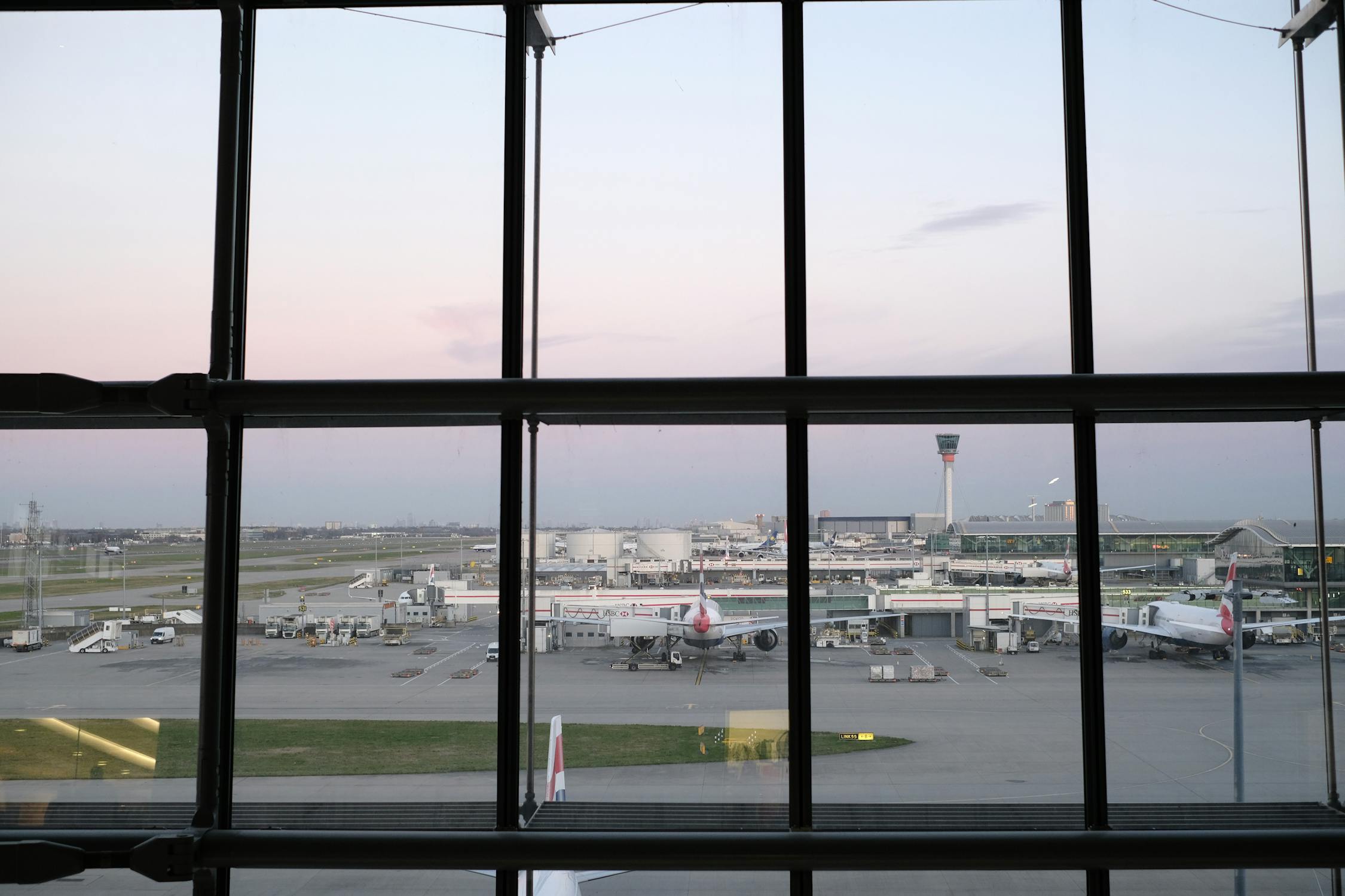 A view of an airport tarmac through large glass windows, showing parked airplanes, an air traffic control tower, and a pastel sky.