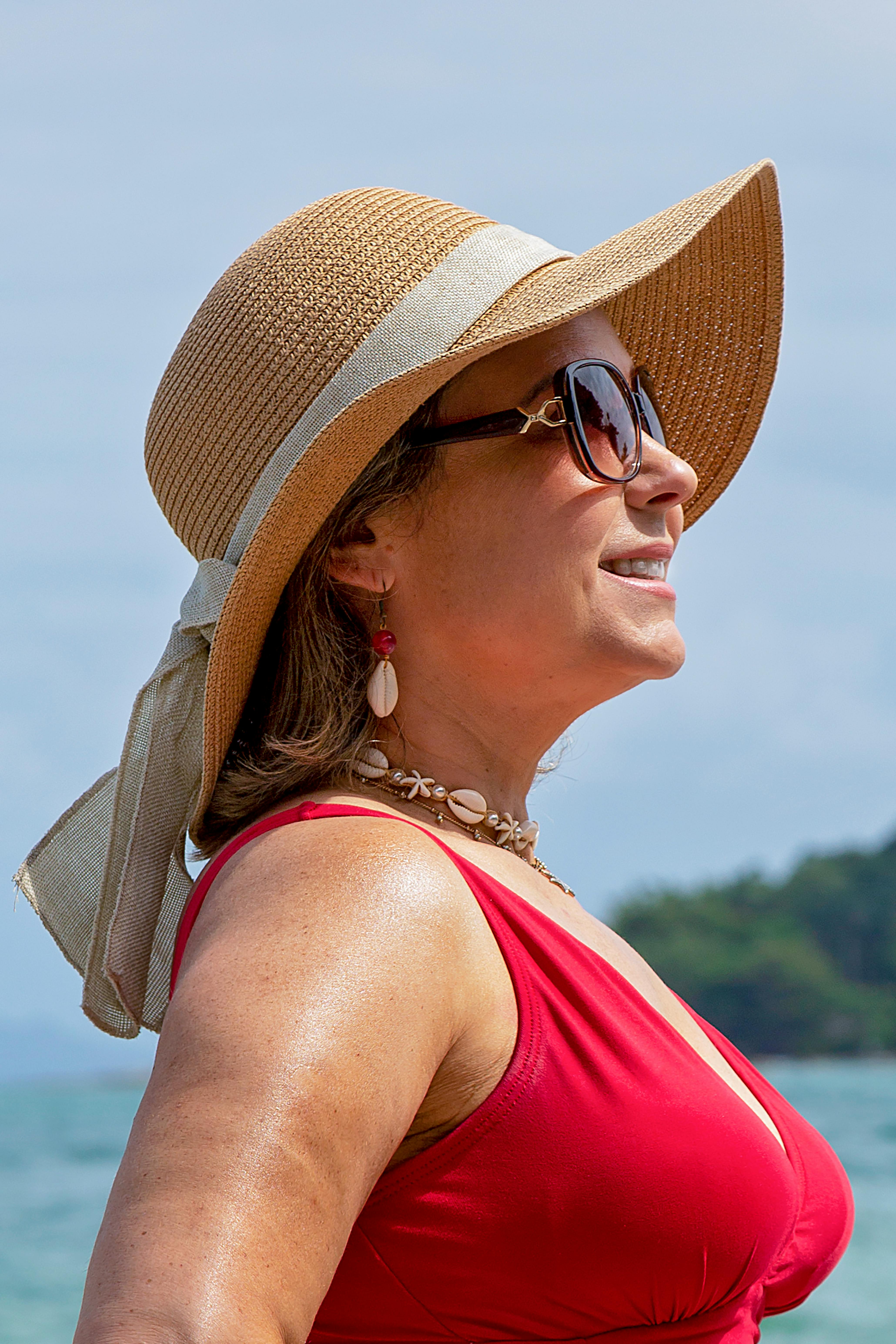 Fashionable woman in sunglasses and straw hat smiling on a sunny beach day, embodying summer vibes.