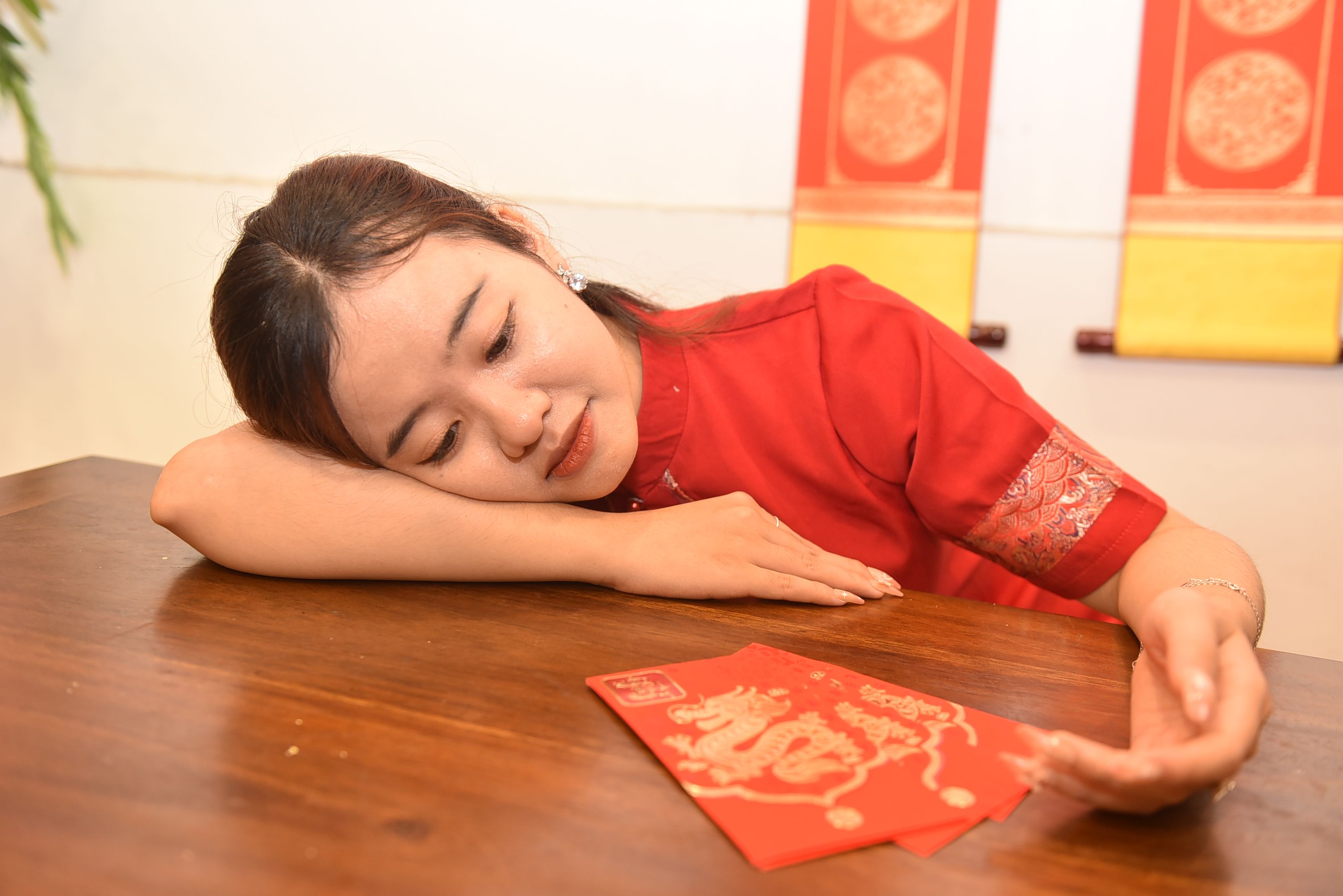 A woman in a red outfit rests her head on a table with festive red envelopes.