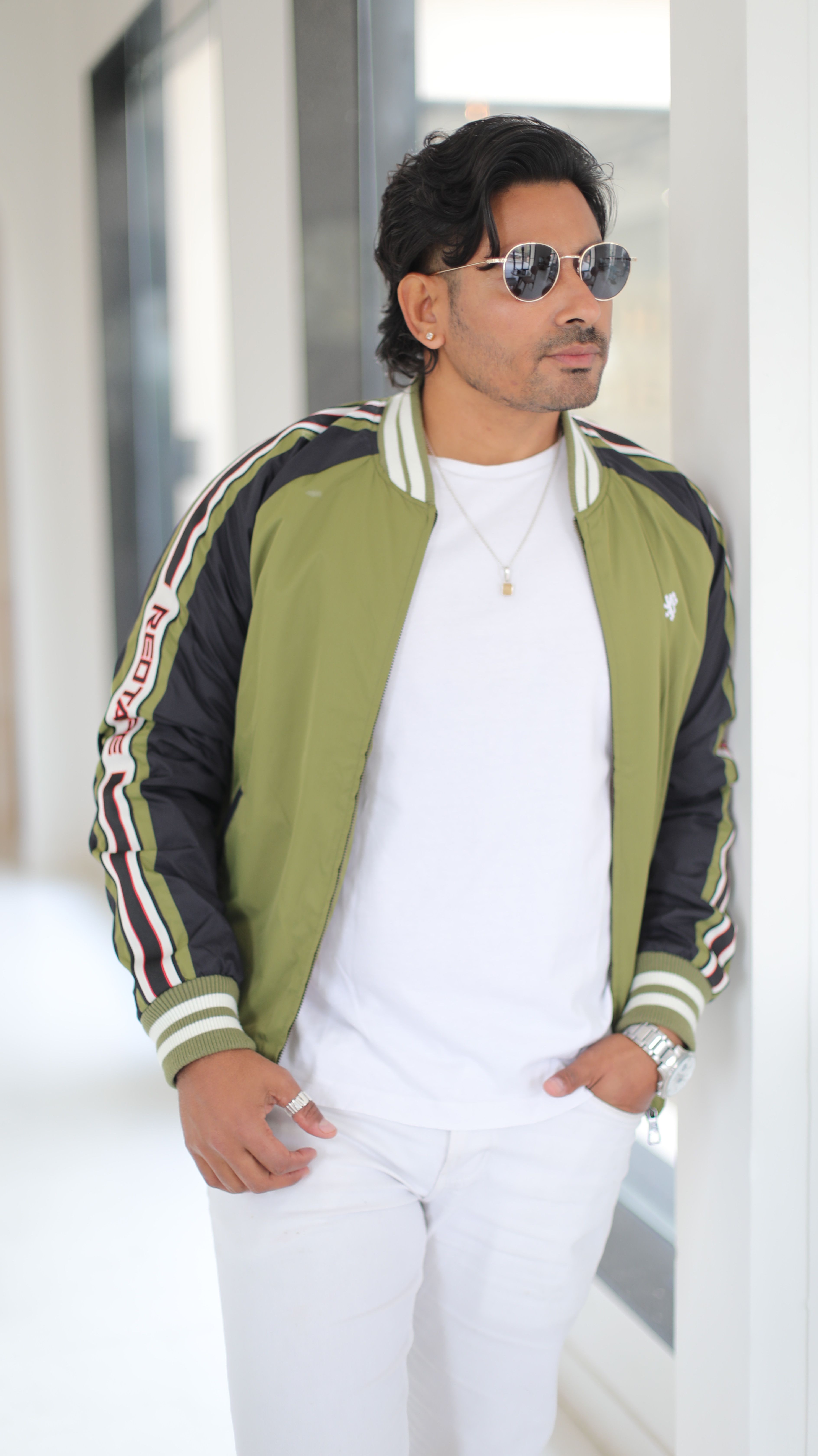 Stylish man in a green jacket and sunglasses standing indoors, exuding confidence and fashion sense.
