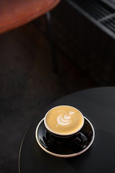 A black coffee cup with intricate latte art sits on a sleek black table in a modern setting.