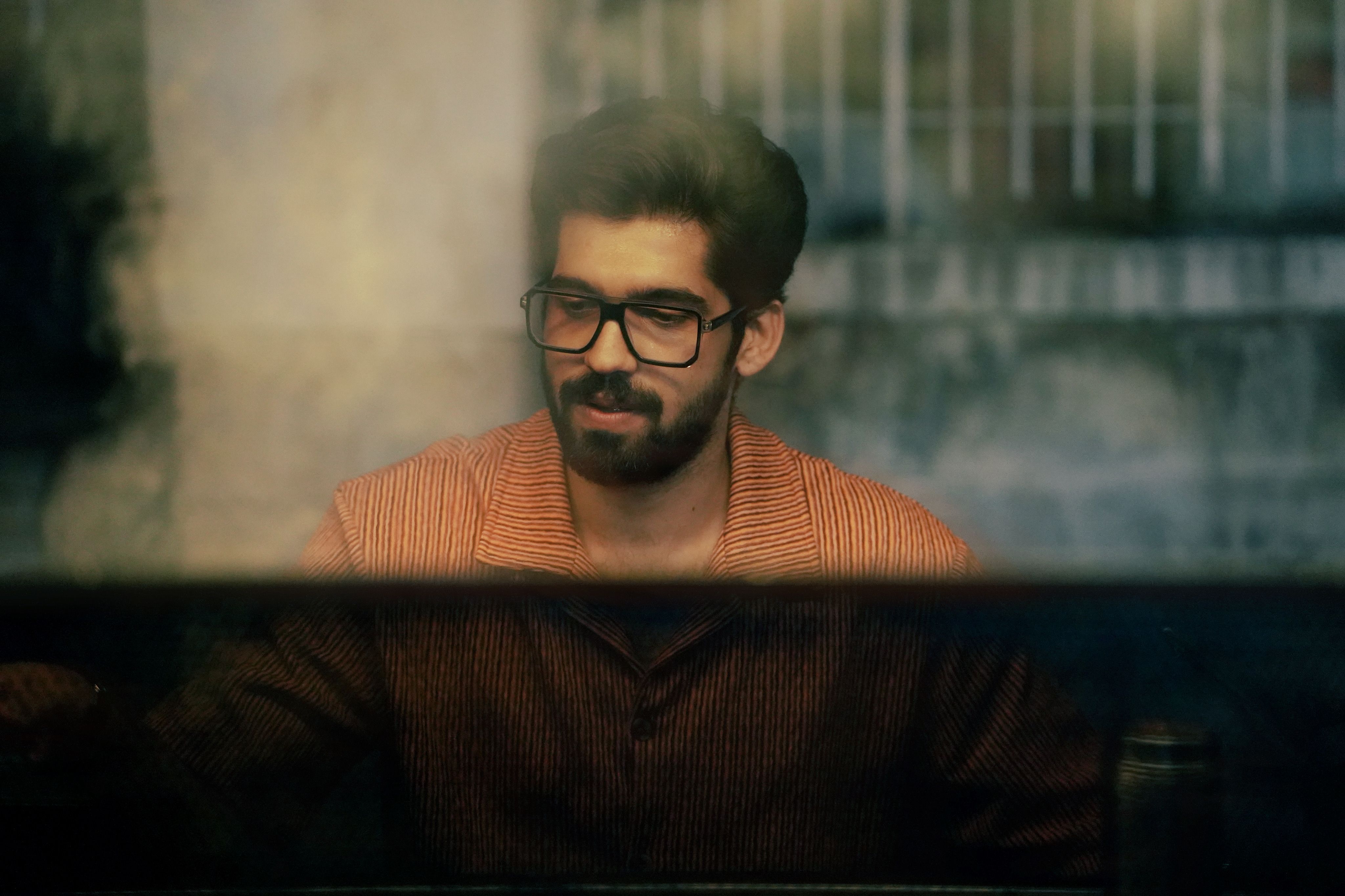Portrait of a bearded man with glasses seen through a window indoors.