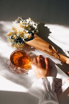 A woman holds a delicate flower bouquet beside a tea cup in warm sunlight on a table.