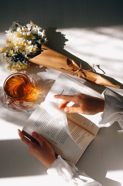Hands holding a book amidst a bouquet and a tea cup in sunlight.