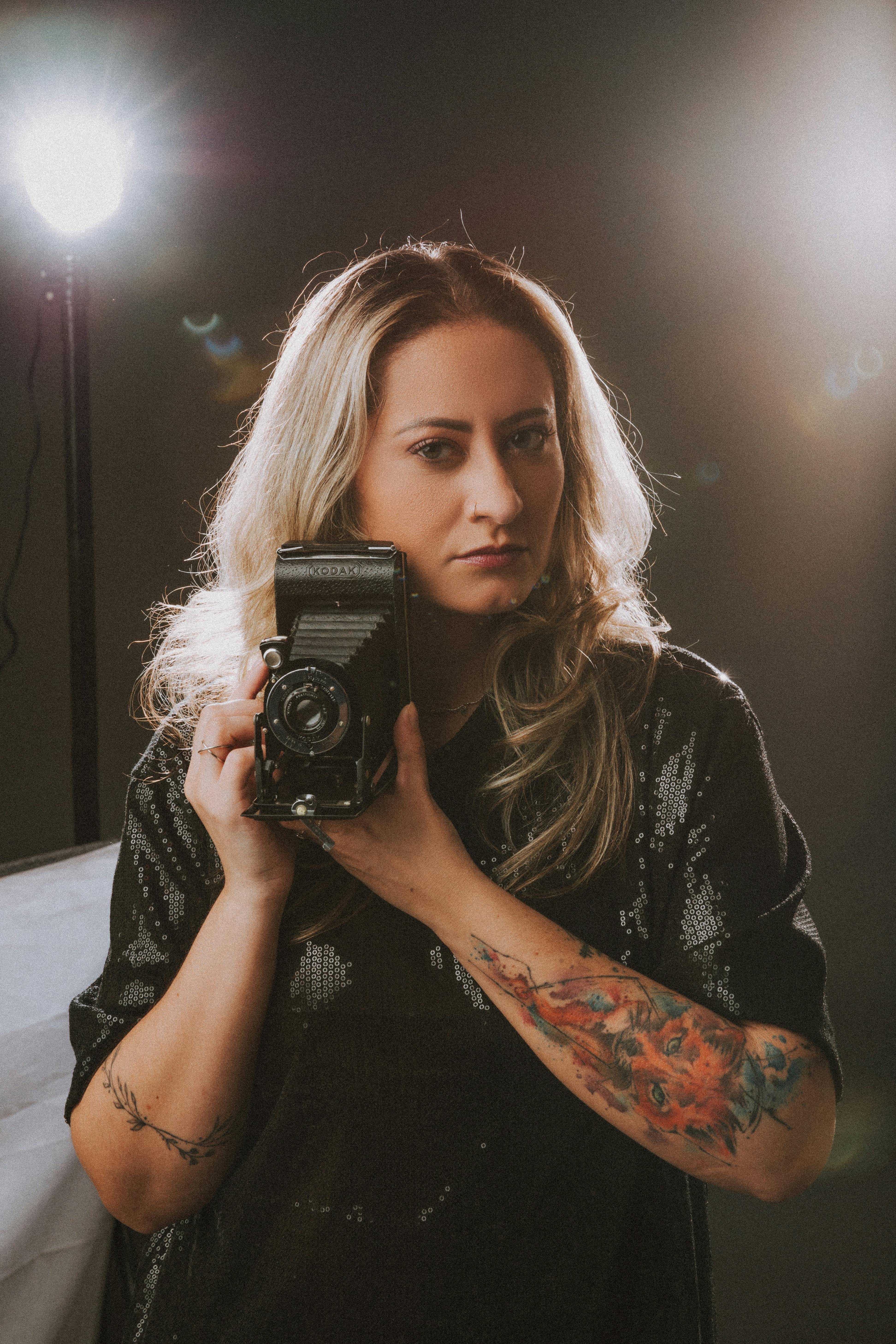 Blonde woman posing with a vintage camera in a studio setting, showcasing tattoos and a serious expression.