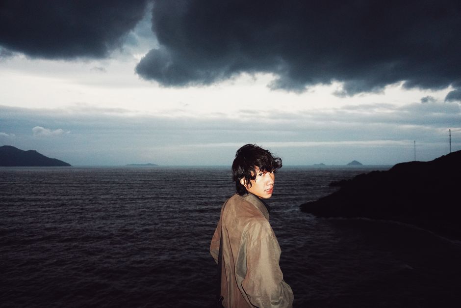 A man with curly hair stands by a rocky coast, gazing at the stormy ocean under dark clouds.