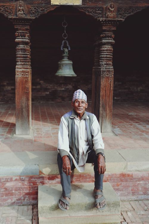 Free Senior man seated on steps in a historic courtyard with carved pillars and a hanging bell. Stock Photo