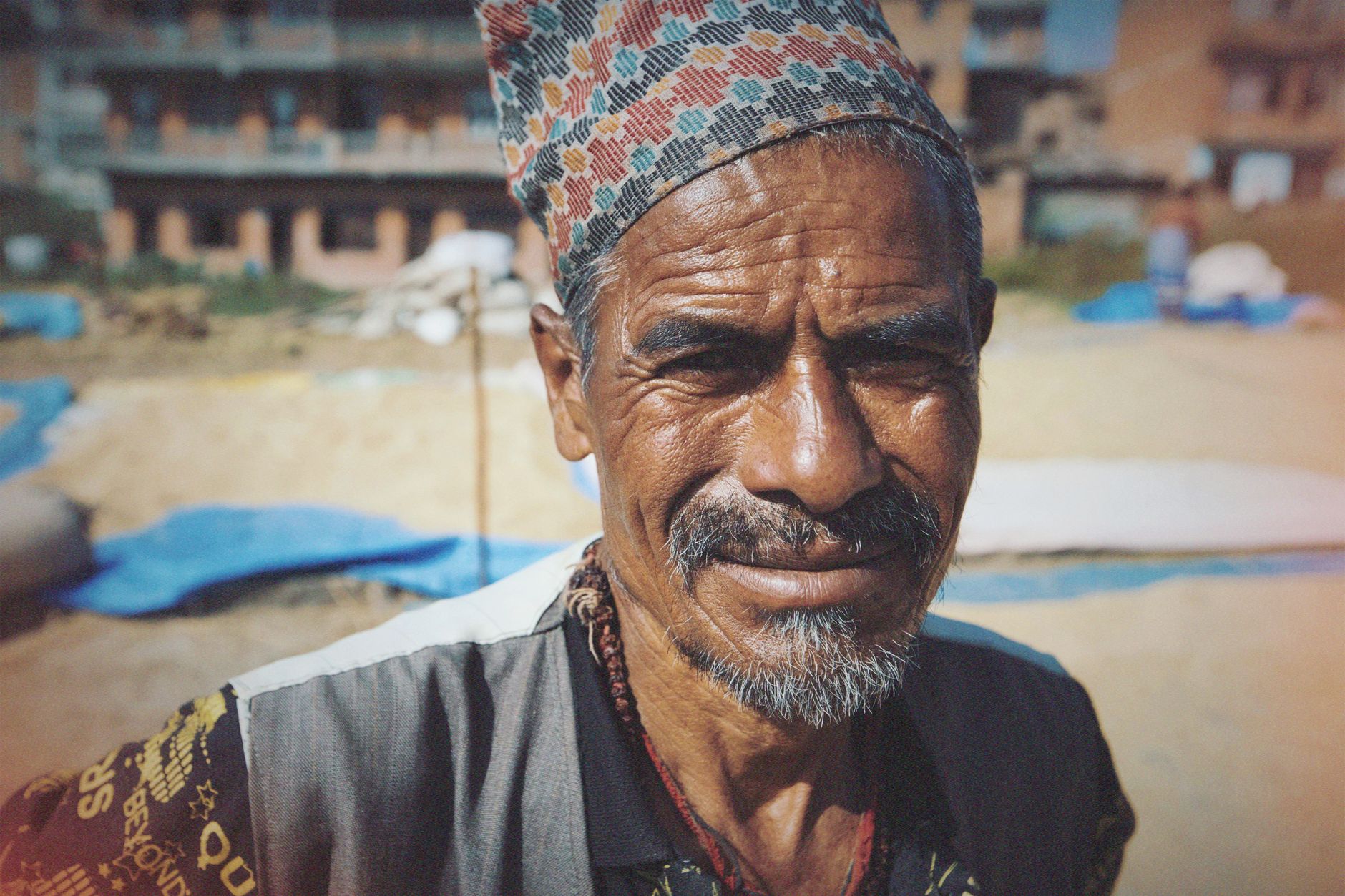 Close-up portrait of a smiling elderly Asian man wearing a patterned cap, outdoors in a sunny setting.
