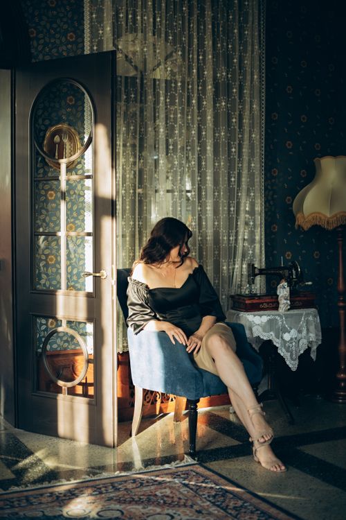 Free Elegant woman sitting in a vintage-style living room with sunlight streaming through the window. Stock Photo