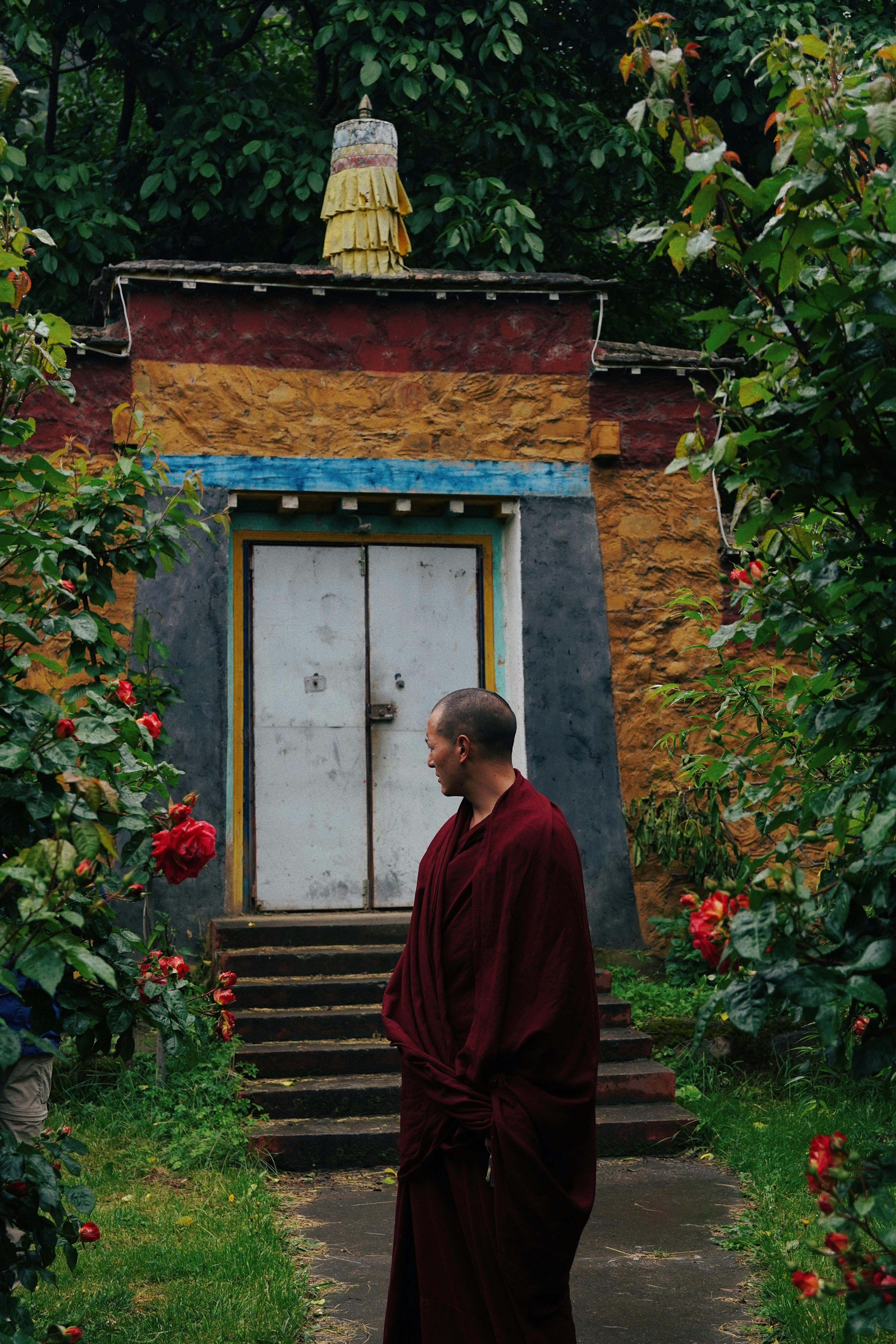 A monk standing near a temple entrance amid lush greenery and red flowers.