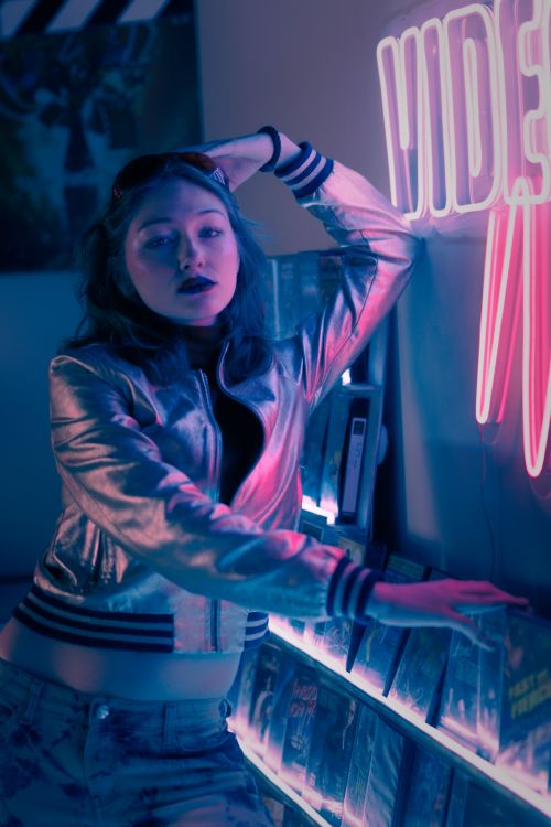 Free Stylish woman in metallic jacket posing with neon lights in a retro video store. Captivating urban portrait. Stock Photo