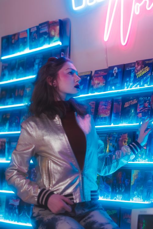 Free Stylish woman in a metallic jacket explores neon-lit album wall, embodying retro aesthetics. Stock Photo