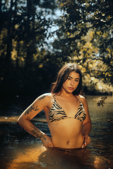 A fashionable woman with tattoos in a zebra print bikini poses confidently in a serene river setting.