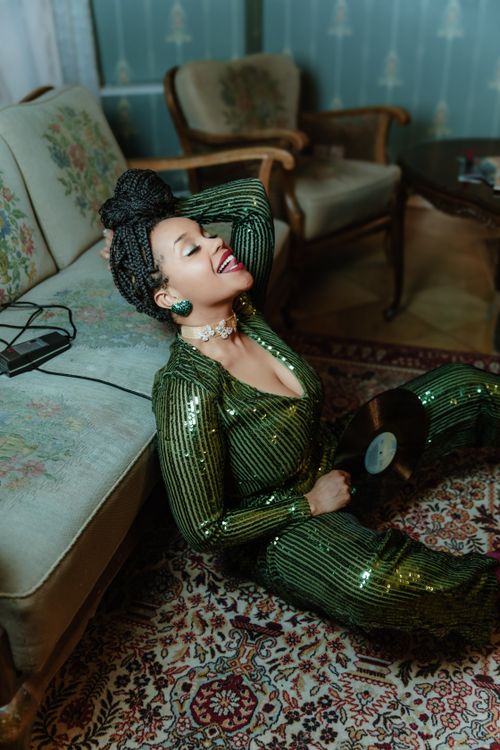 Free Stylish woman in a sparkly green outfit enjoying music indoors, sitting on carpet. Stock Photo