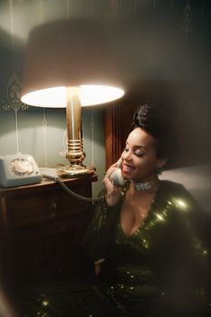 A stylish woman in a green dress smiles while talking on a vintage rotary phone beside a golden lamp.