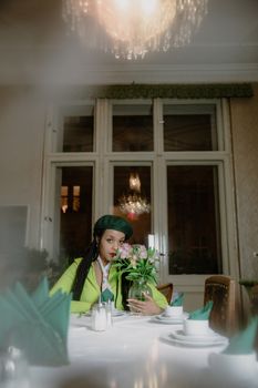 Stylish woman with braided hair and beret dining elegantly with flowers indoors.