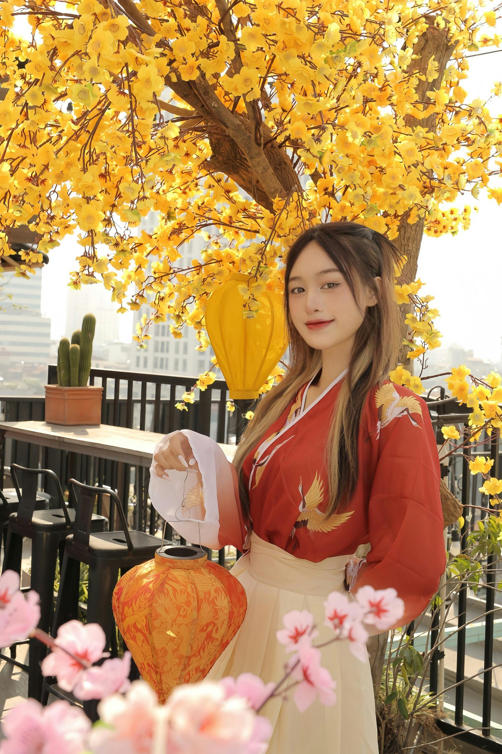 Asian woman in red blouse and beige skirt posing with lantern under blooming yellow tree.