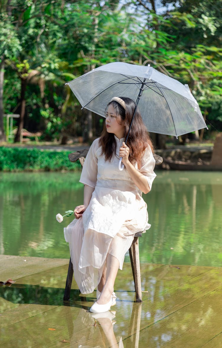 Woman In White Dress Sitting With Umbrella On Chair On Water