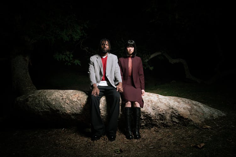 Woman And Man In Suits Sitting On Tree Trunk At Night