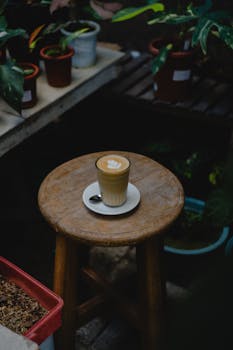 A latte with intricate art sits on a wooden stool surrounded by potted plants in a rustic coffee shop setting.