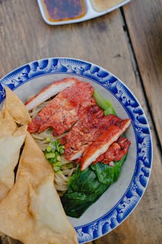 Delicious Indonesian chicken noodle dish with vegetables, fried wontons, and sauces in a blue rimmed bowl.