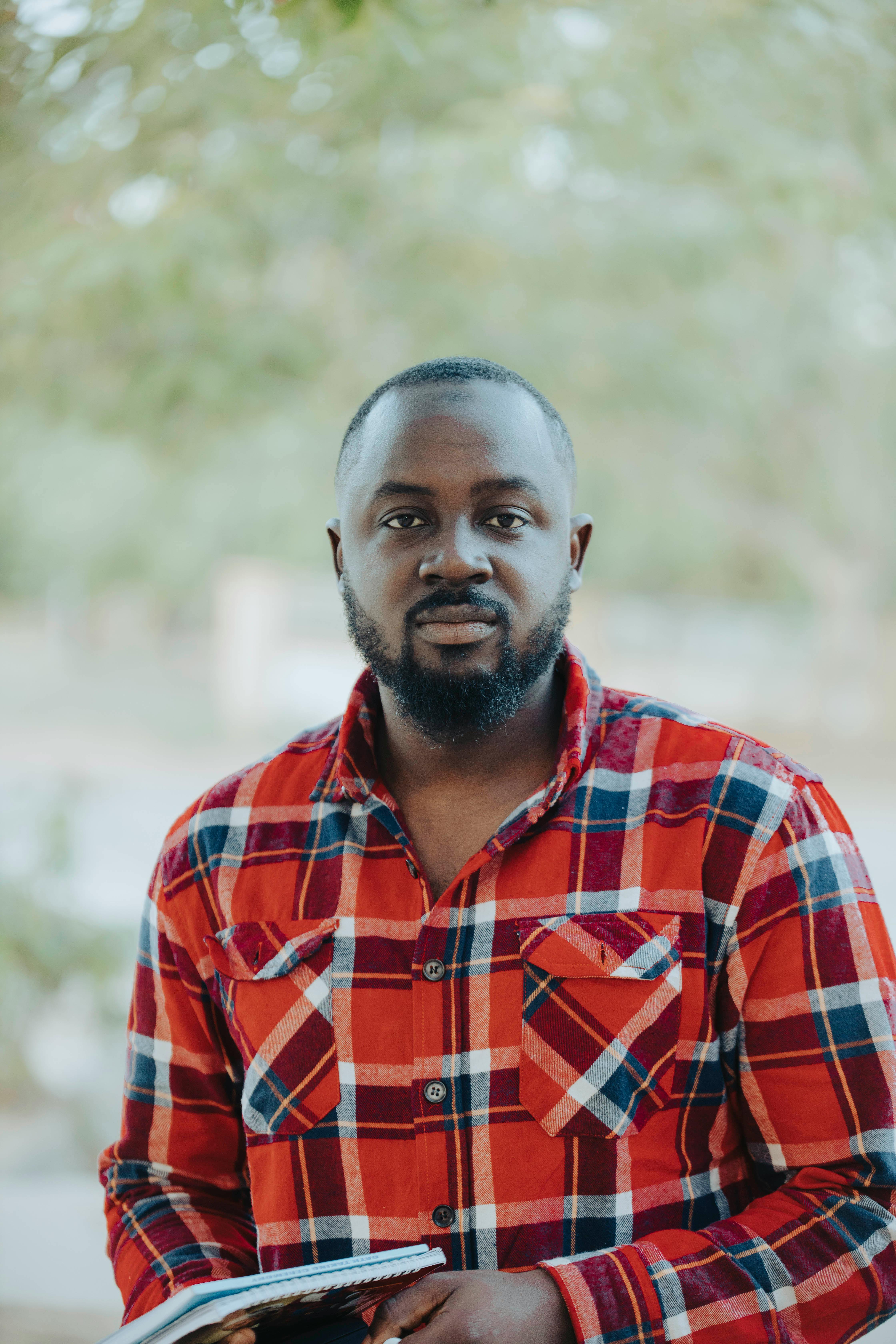 Portrait of a bearded man in a red plaid shirt, standing outdoors with a confident look.