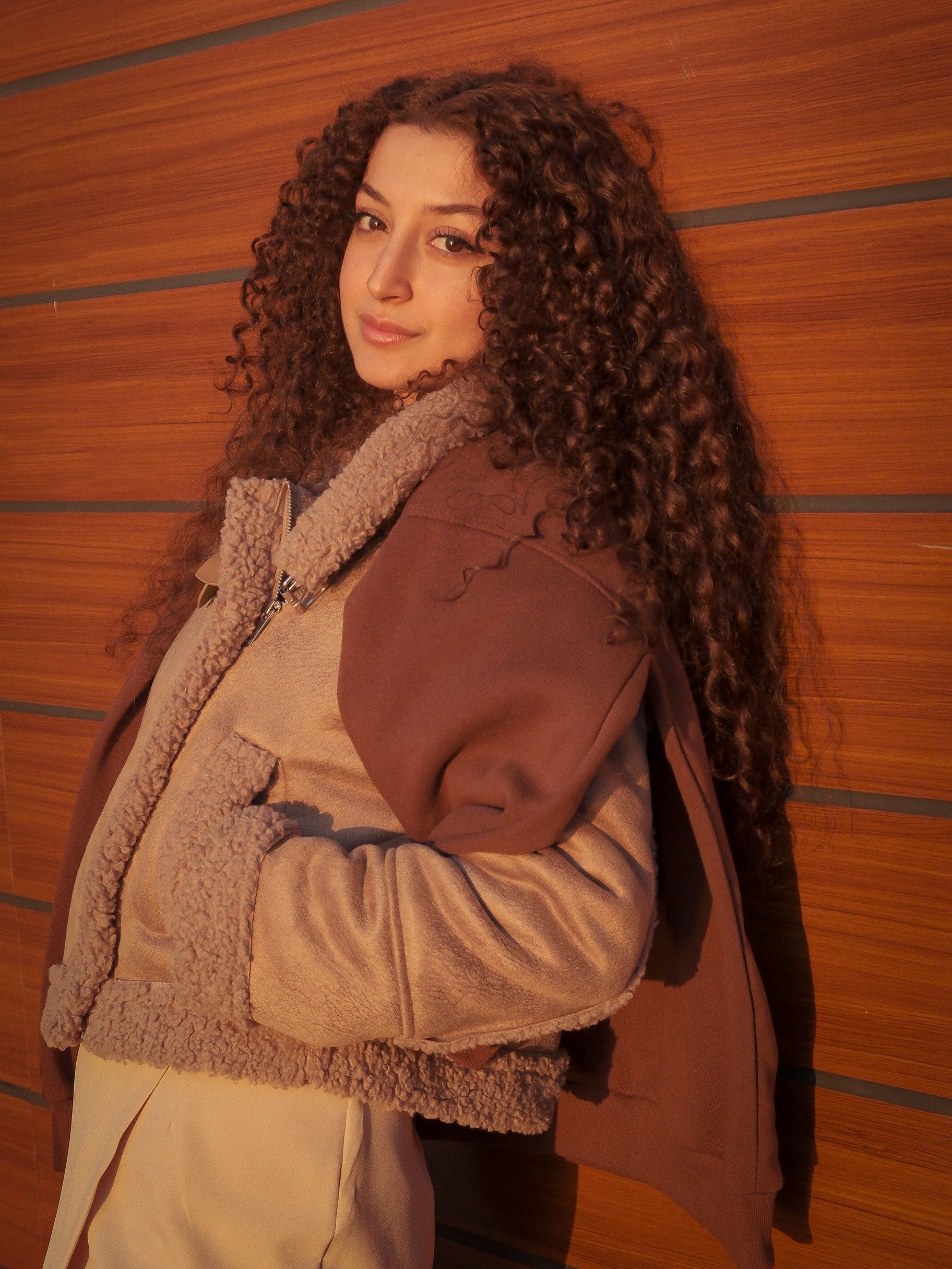 Fashionable woman with curly hair wearing a winter coat, leaning against a wooden wall in warm light.