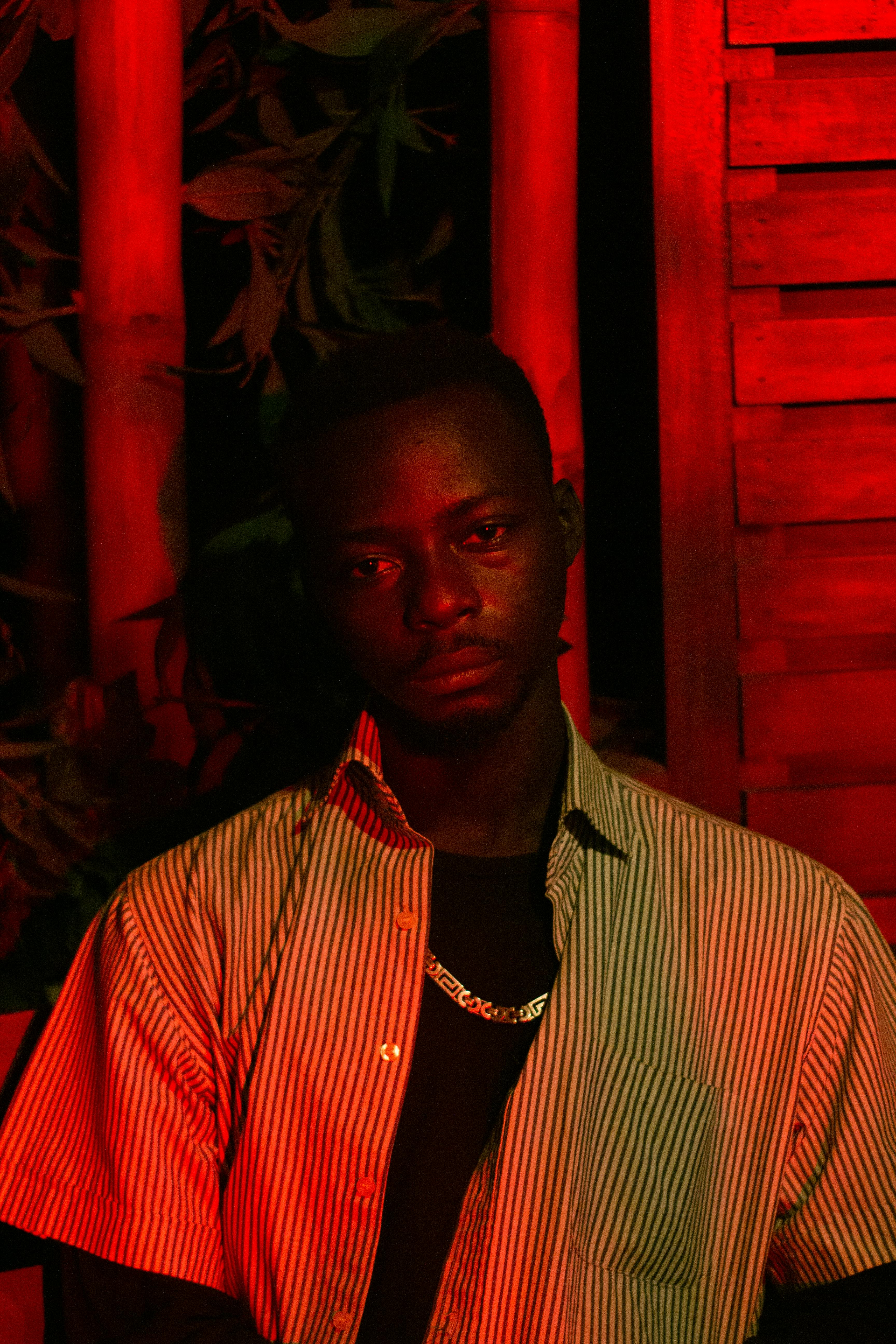 Artistic portrait of a man in a striped shirt and necklace, bathed in dramatic red lighting.