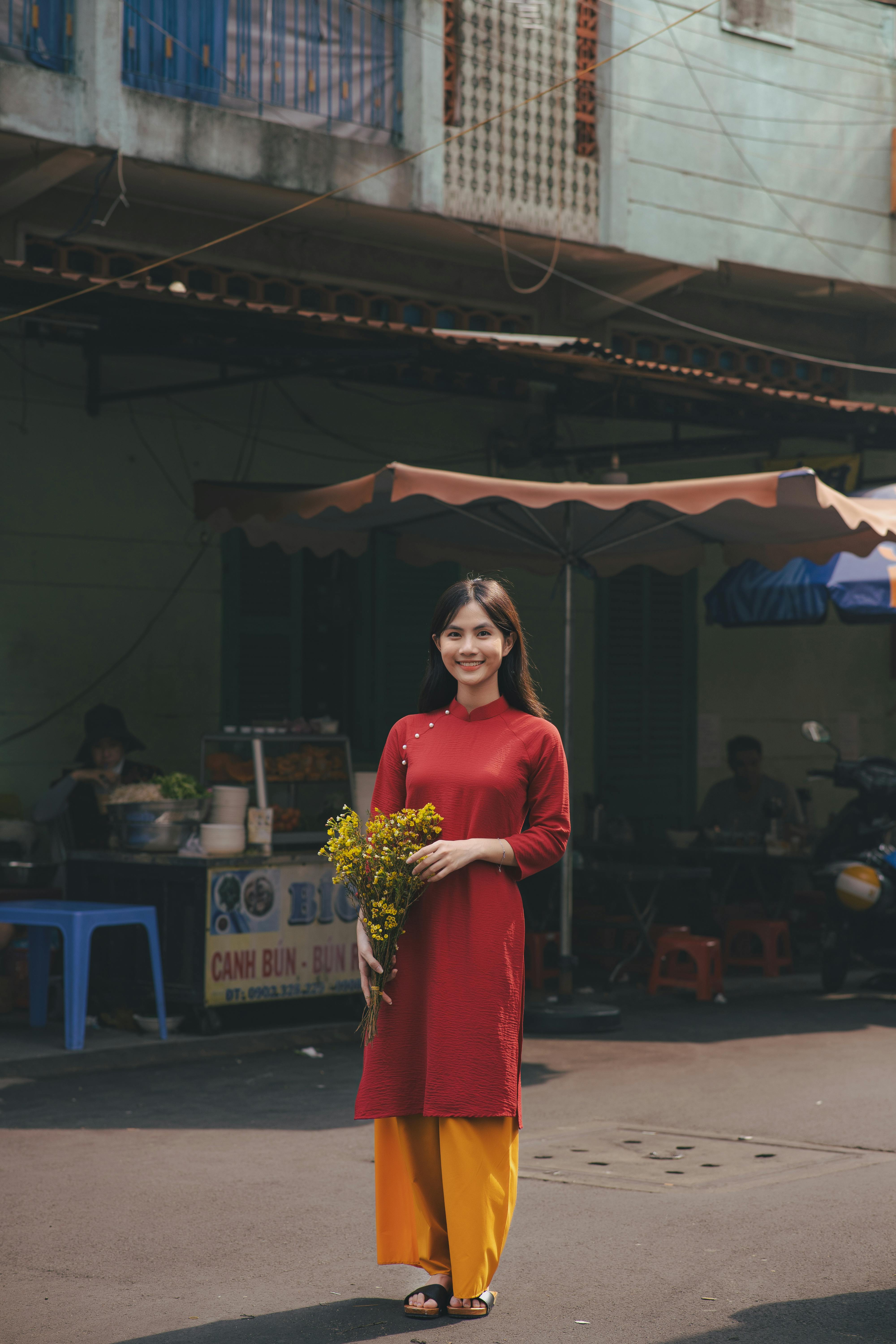 Free Smiling Asian woman in traditional red dress holding flowers in urban setting. Stock Photo